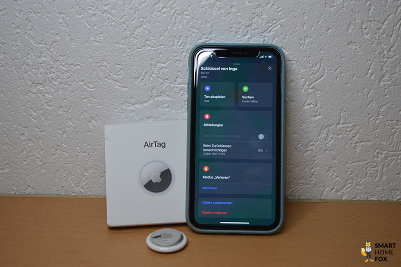 iPhone mit Wo ist App, AirTag und Verpackung sind vor einer weißen Wand zu sehen.